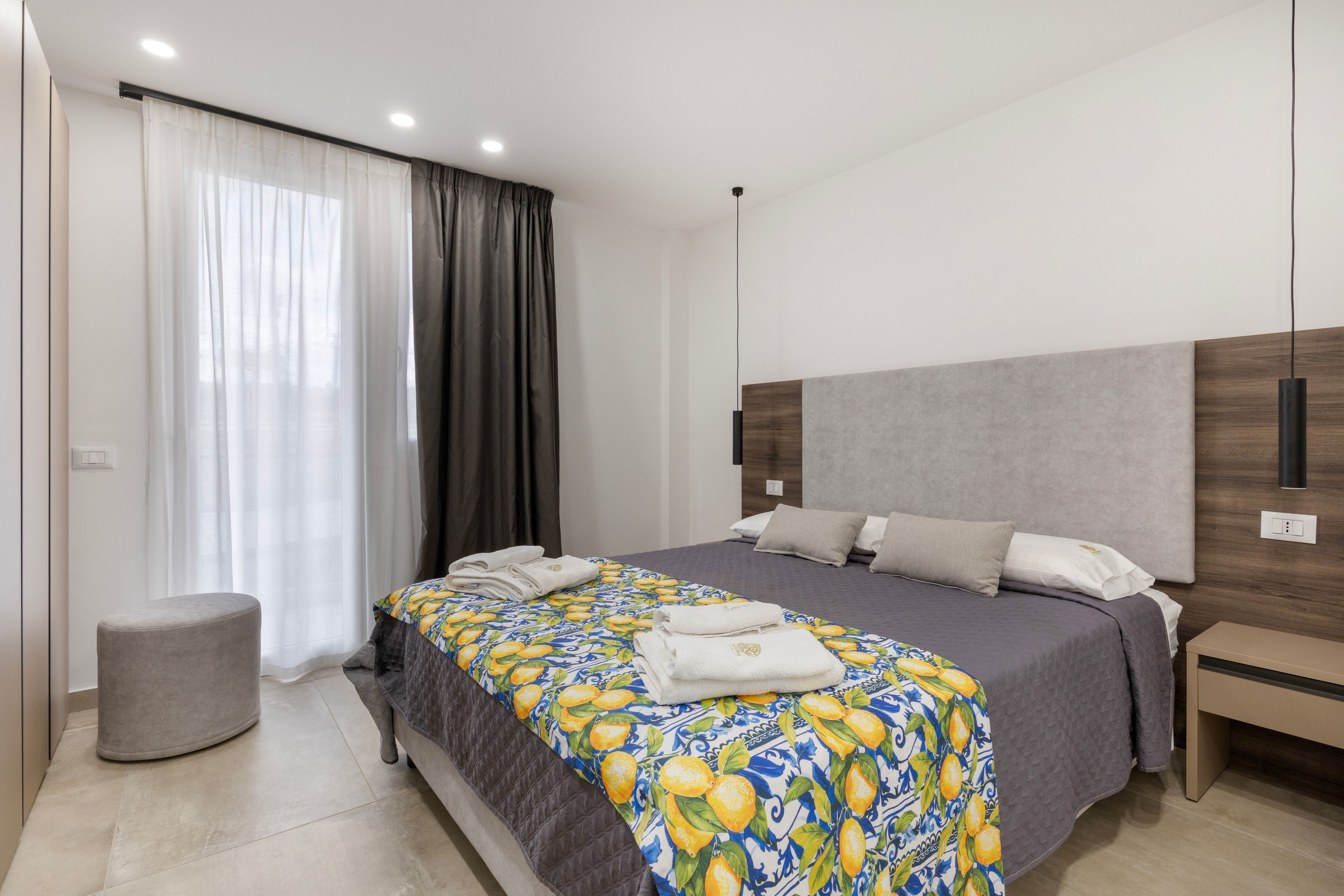 A modern bedroom with a large bed, a bedside table, a small stool, and a window with curtains.