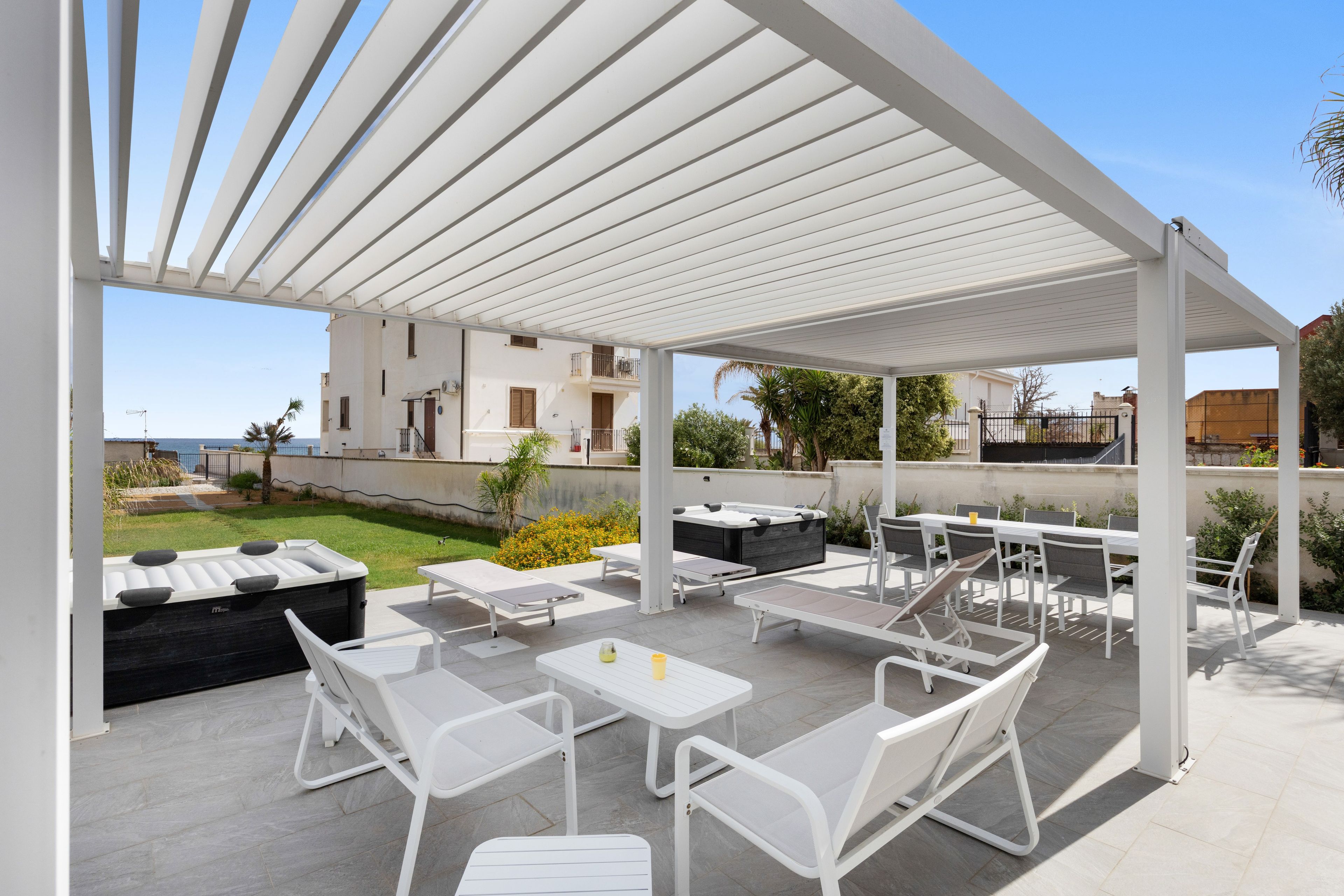 An outdoor patio with white furniture, including lounge chairs and tables, under a white pergola.