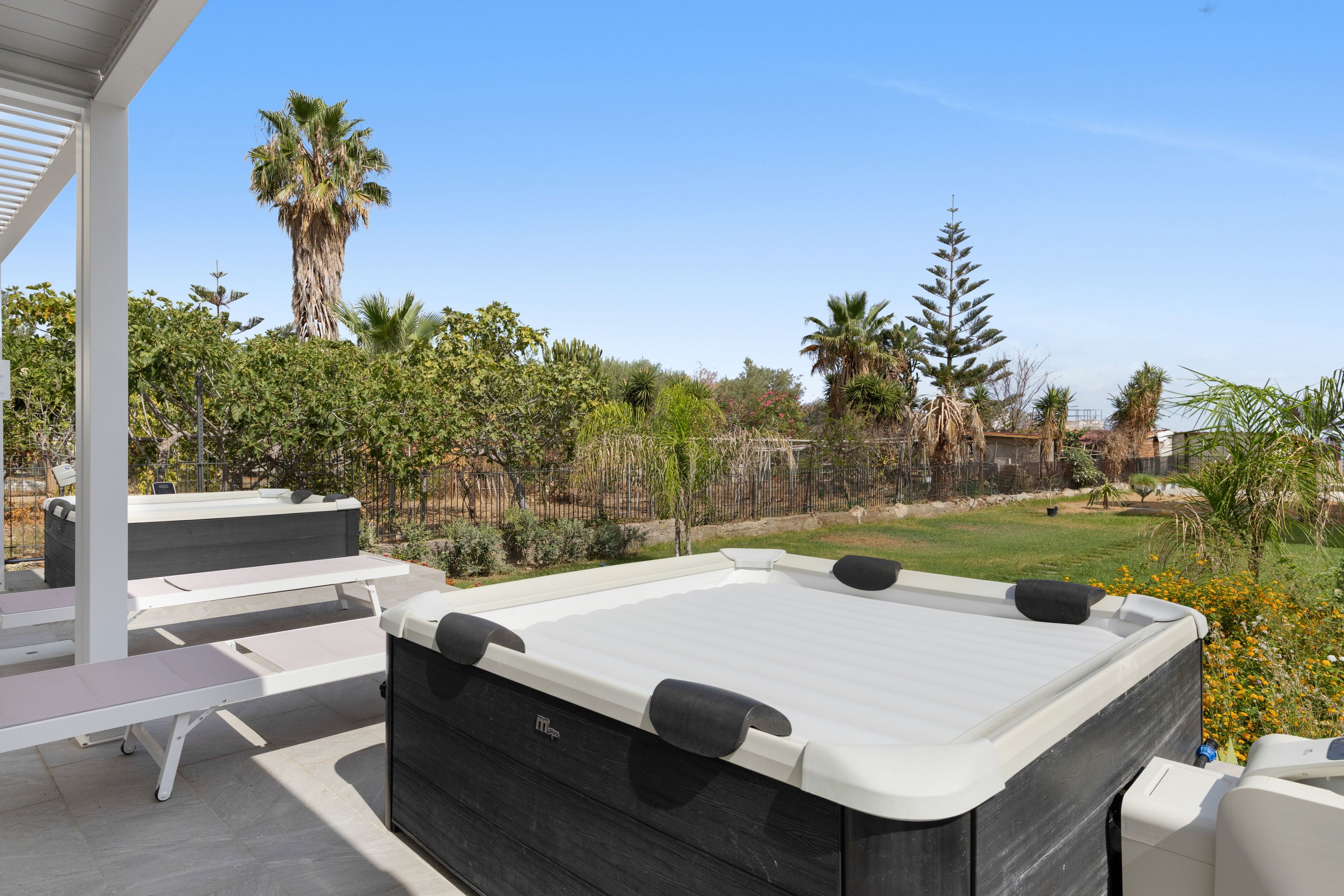 Uma banheira de hidromassagem com exterior branco e preto, colocada num pátio com vista para um jardim ajardinado e um céu azul límpido.