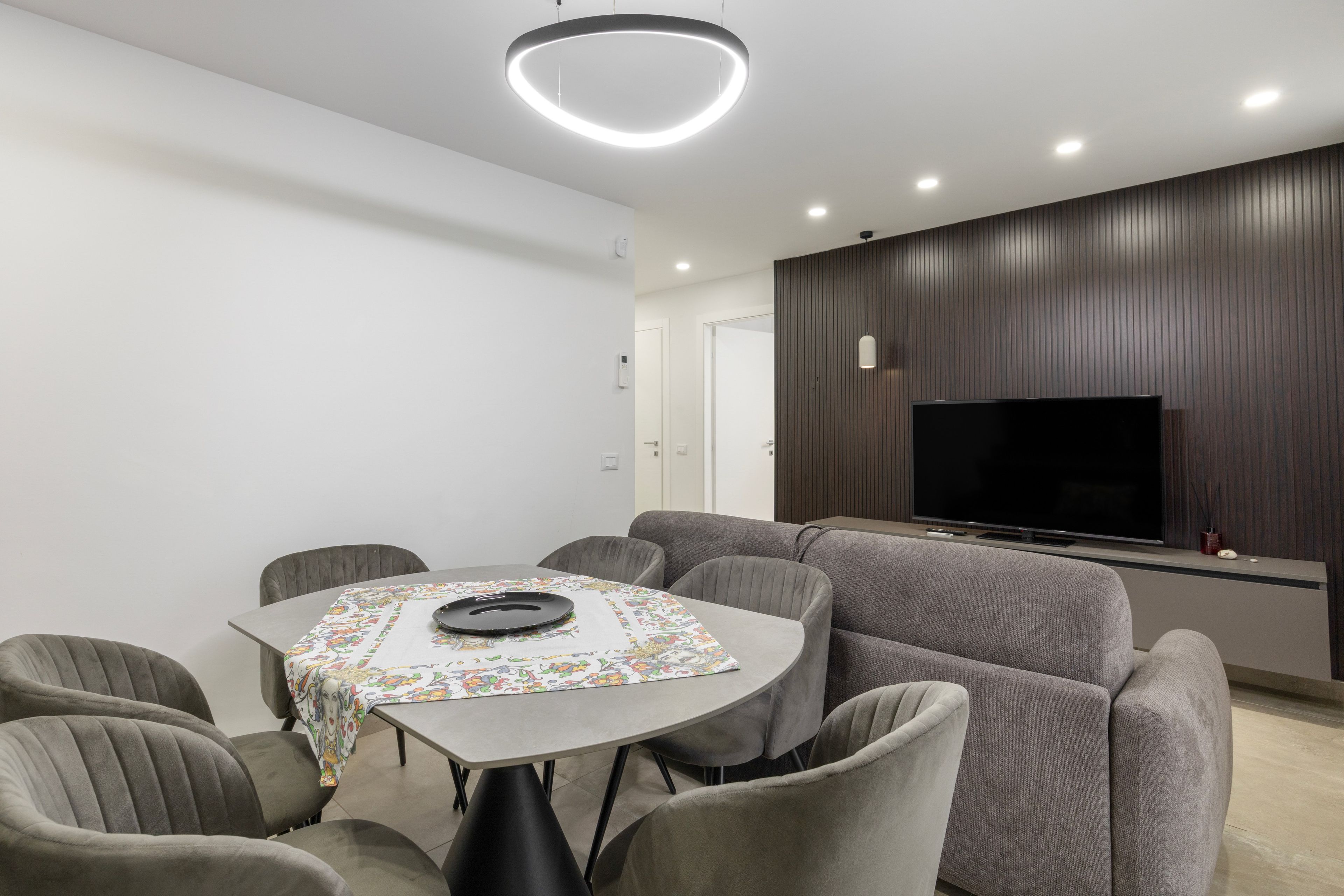 A modern living room with a round table, grey sofas, a flat-screen TV, and a ceiling light fixture.