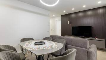 A modern living room with a round table, grey sofas, a flat-screen TV, and a ceiling light fixture.