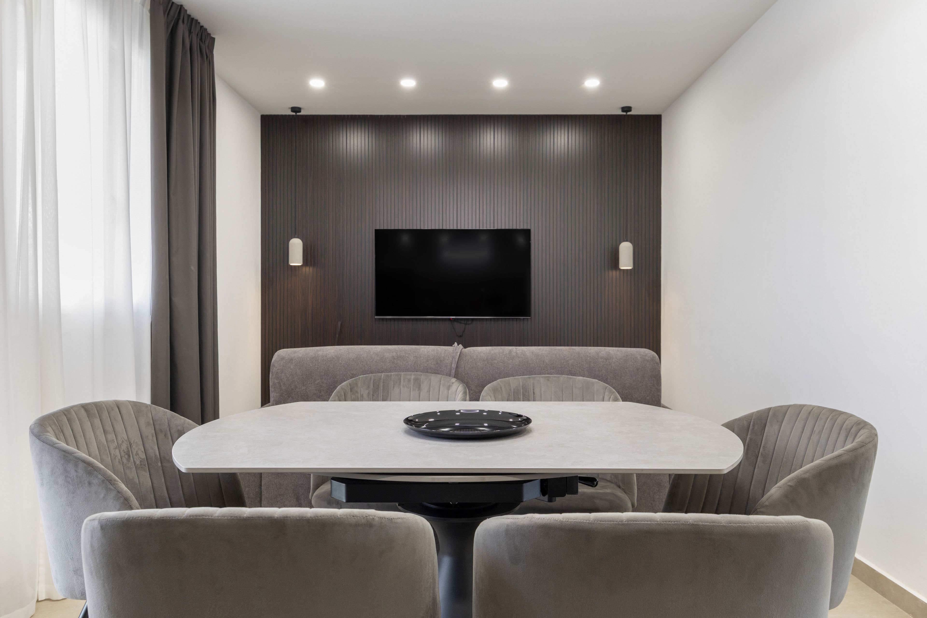 Una moderna sala da pranzo con un tavolo rotondo, sei sedie imbottite, un televisore a schermo piatto montato a parete e illuminazione a incasso nel soffitto.