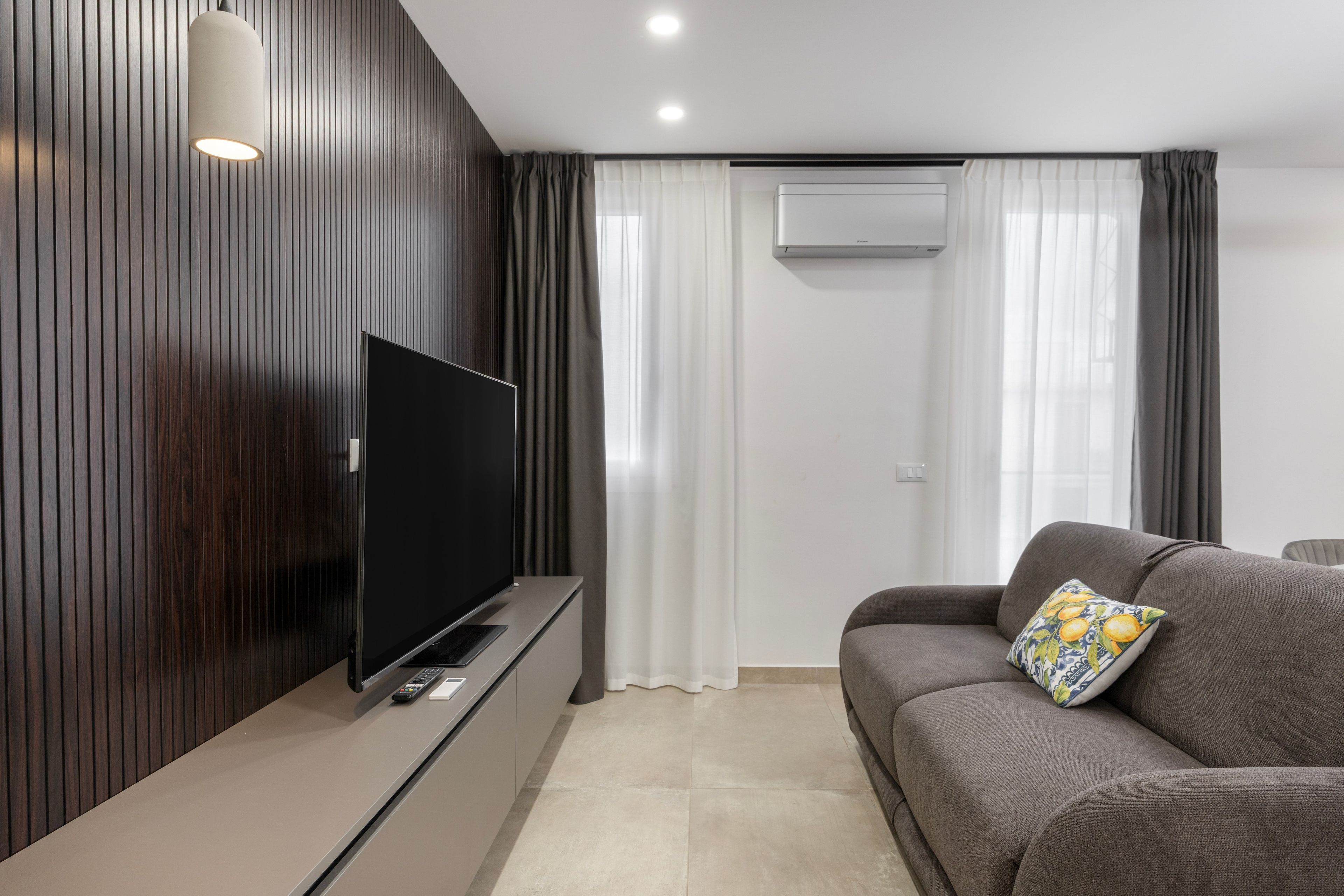 A modern living room with a flat-screen TV mounted on a wooden panel wall, a grey sofa with a decorative pillow, and a white air conditioning unit on the wall.