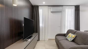 A modern living room with a flat-screen TV mounted on a wooden panel wall, a grey sofa with a decorative pillow, and a white air conditioning unit on the wall.