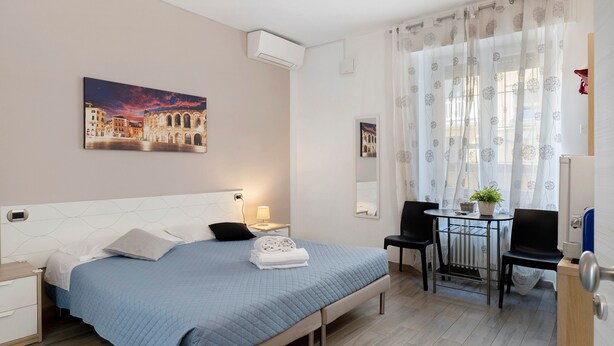 Une chambre d’hôtel comprenant un lit, une table de chevet, une lampe, une chaise, une table avec une plante et une fenêtre avec des rideaux.
