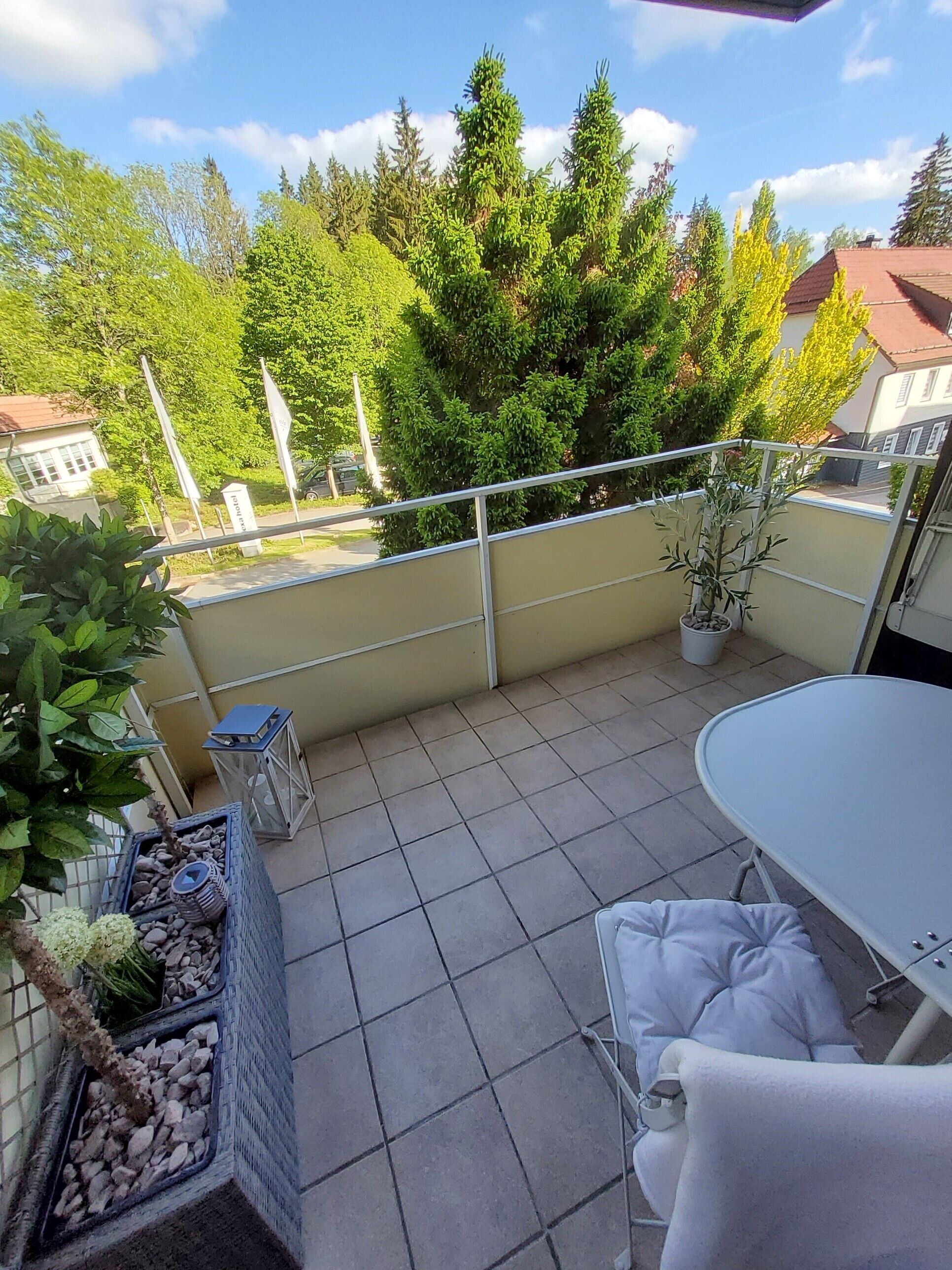 A balcony with a table and chairs, potted plants, and a view of trees and buildings.