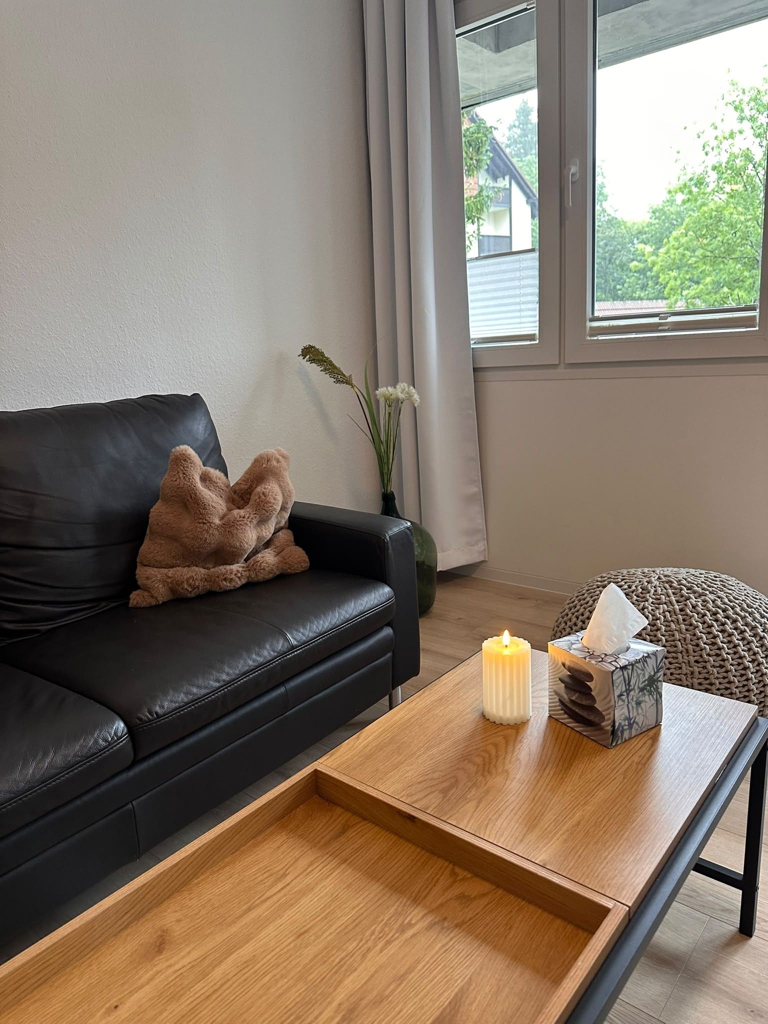 Una sala de estar moderna con un sofá de cuero negro, una mesa de centro de madera con una vela, y vistas al verdor exterior.