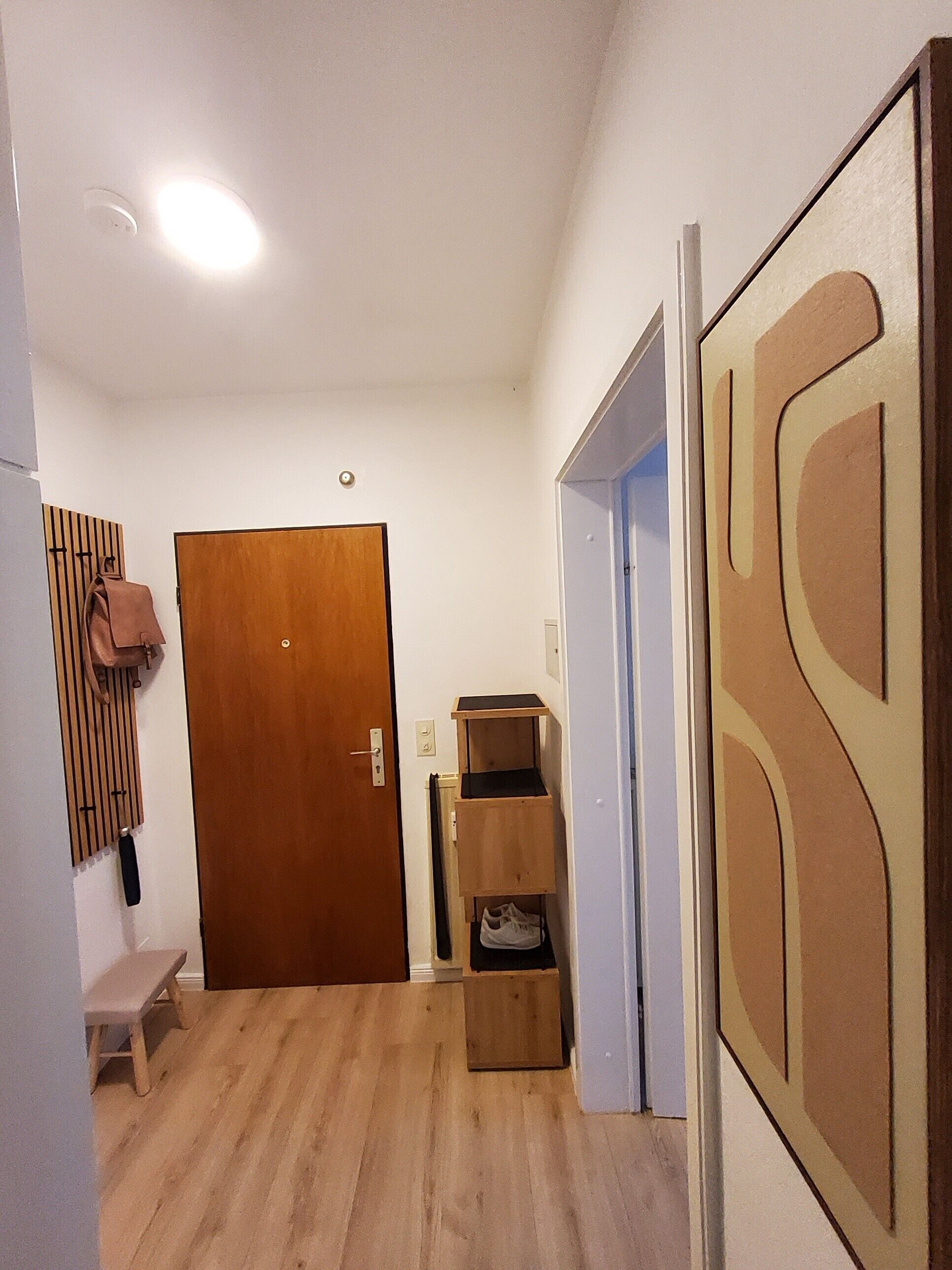 Ein Korridor mit einer Holztür, einer Sitzbank, einem Schuhregal und einer Wand mit geometrischem Muster.