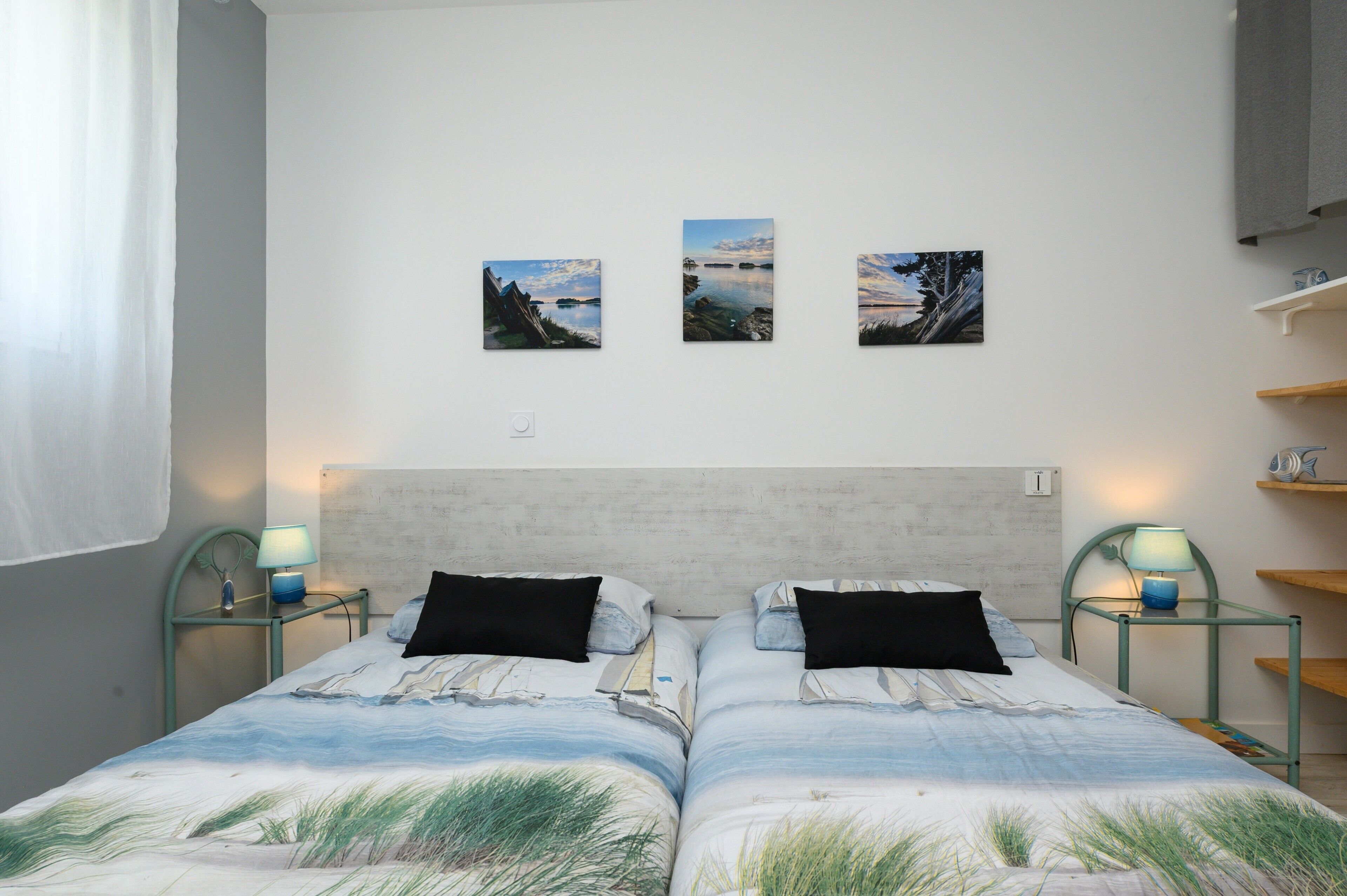 A double bed with a coastal-themed bedspread, two bedside tables with lamps, and three framed landscape paintings on the wall.