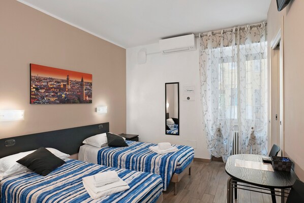 A hotel room with two beds, a cityscape painting, a mirror, a table, and a window with curtains.