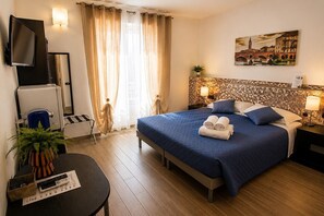 A hotel room with a bed, a TV, a desk with a plant, and a painting on the wall.