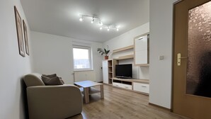 A modern living room with a sofa, a TV on a wooden stand, a small table, and a window with a view. - Apartment "New Holiday Home for 2 Guests" with Shared Garden, Balcony & Wi-Fi (Oelsnitz/Erzgebirge)