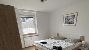 A bedroom with a bed, a window, a radiator, a framed picture, and a bedside table. - Apartment "New Holiday Home for 2 Guests" with Shared Garden, Balcony & Wi-Fi (Oelsnitz/Erzgebirge)
