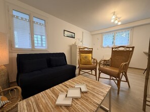 A room with a black sofa, two wicker chairs, a wooden coffee table, and a wall-mounted light fixture. - Appartement des Camélias – 100m from the Beach (La Tranche-sur-Mer)