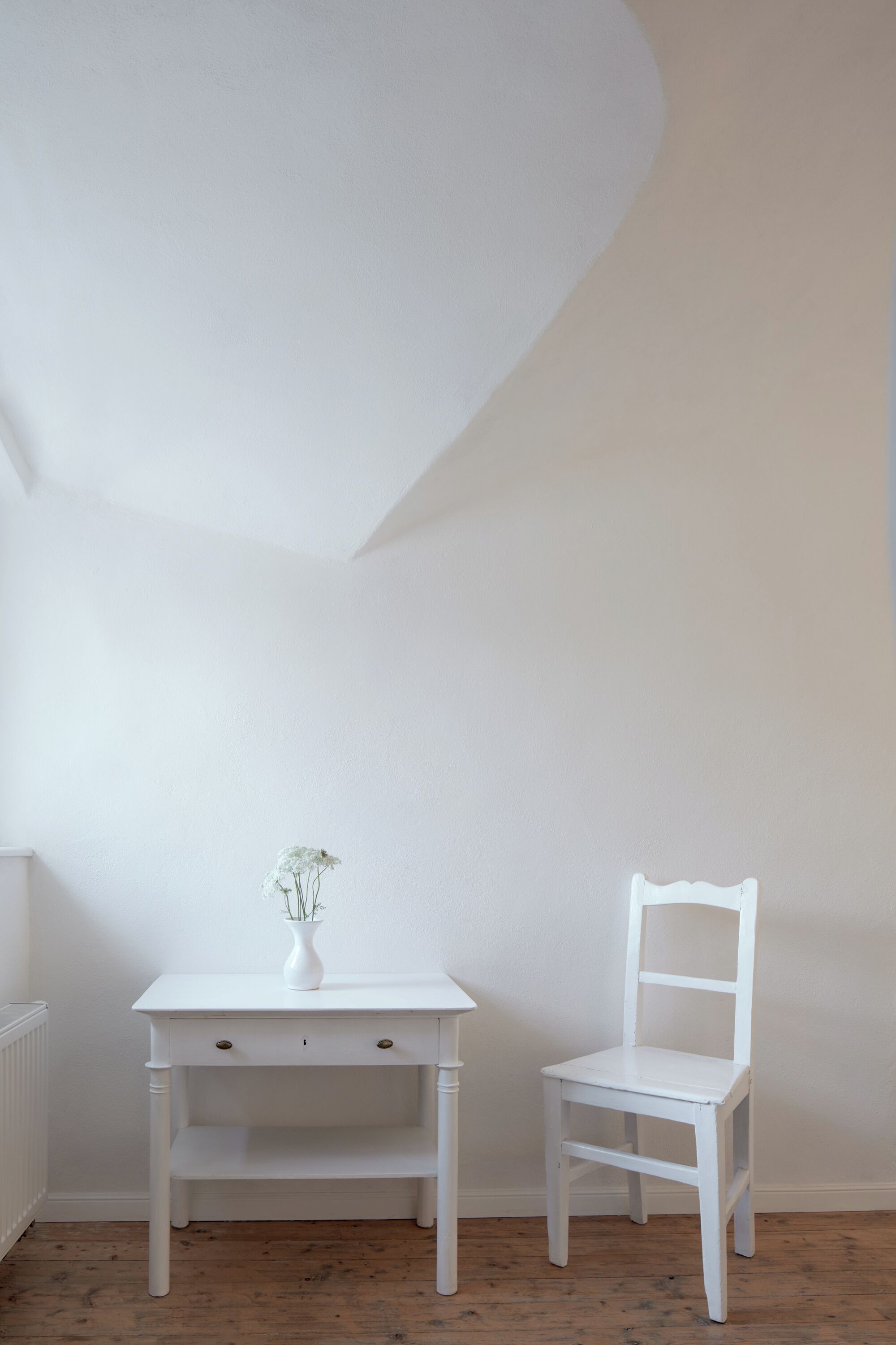 A white table with a vase and a chair in a room with white walls and wooden flooring.