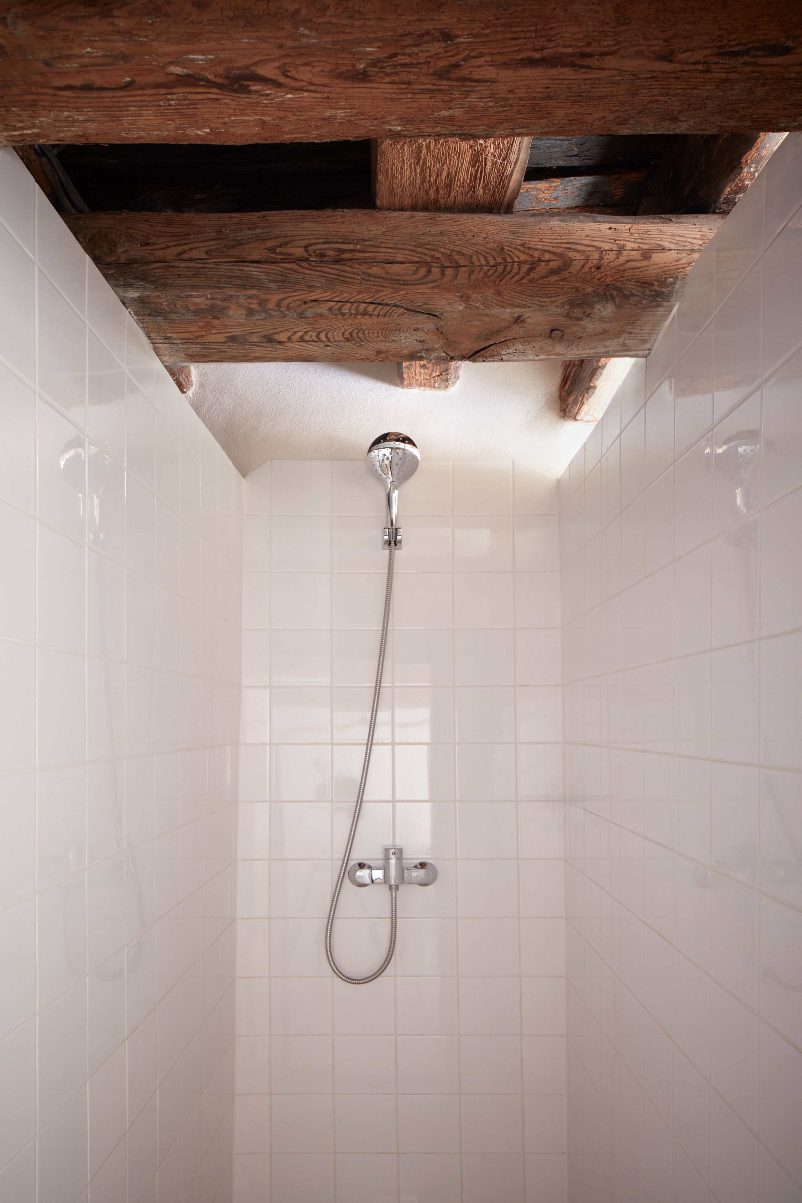 A shower with a wooden ceiling beam, white tiled walls, and a chrome showerhead.