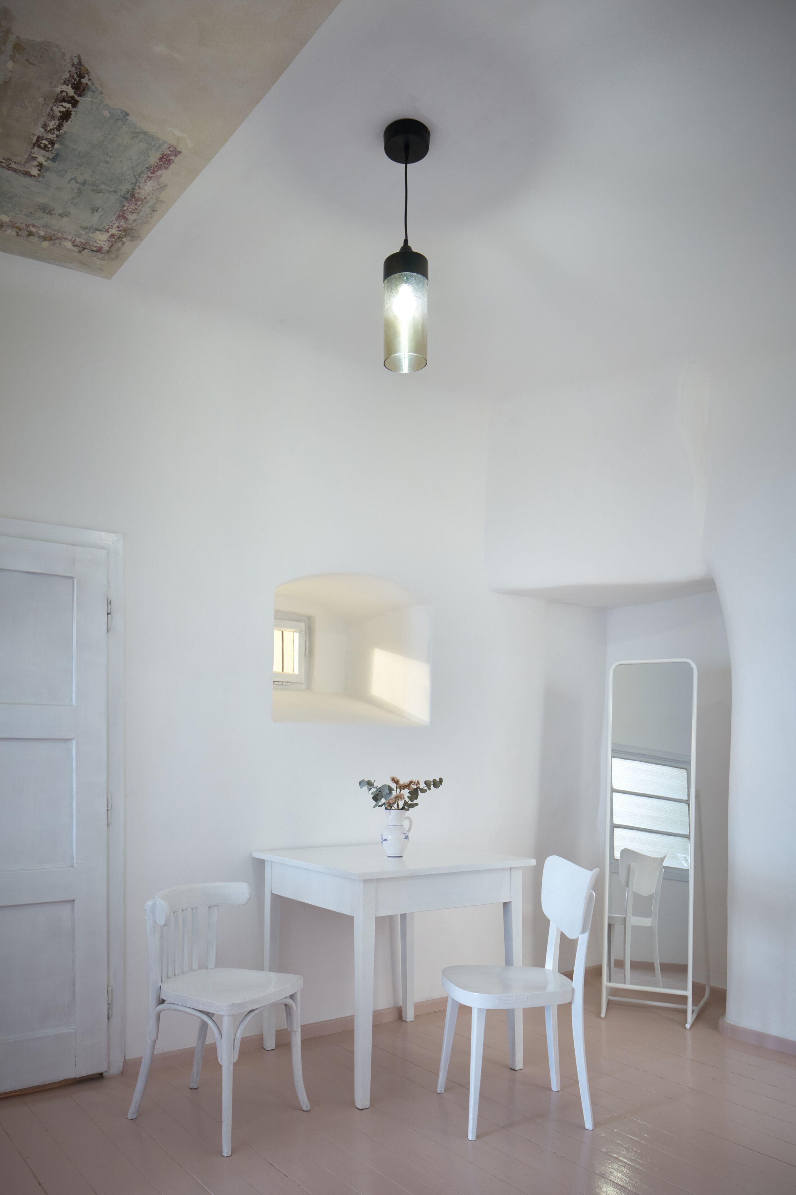 A room with a white table, two white chairs, a hanging light fixture, a mirror, and a door.