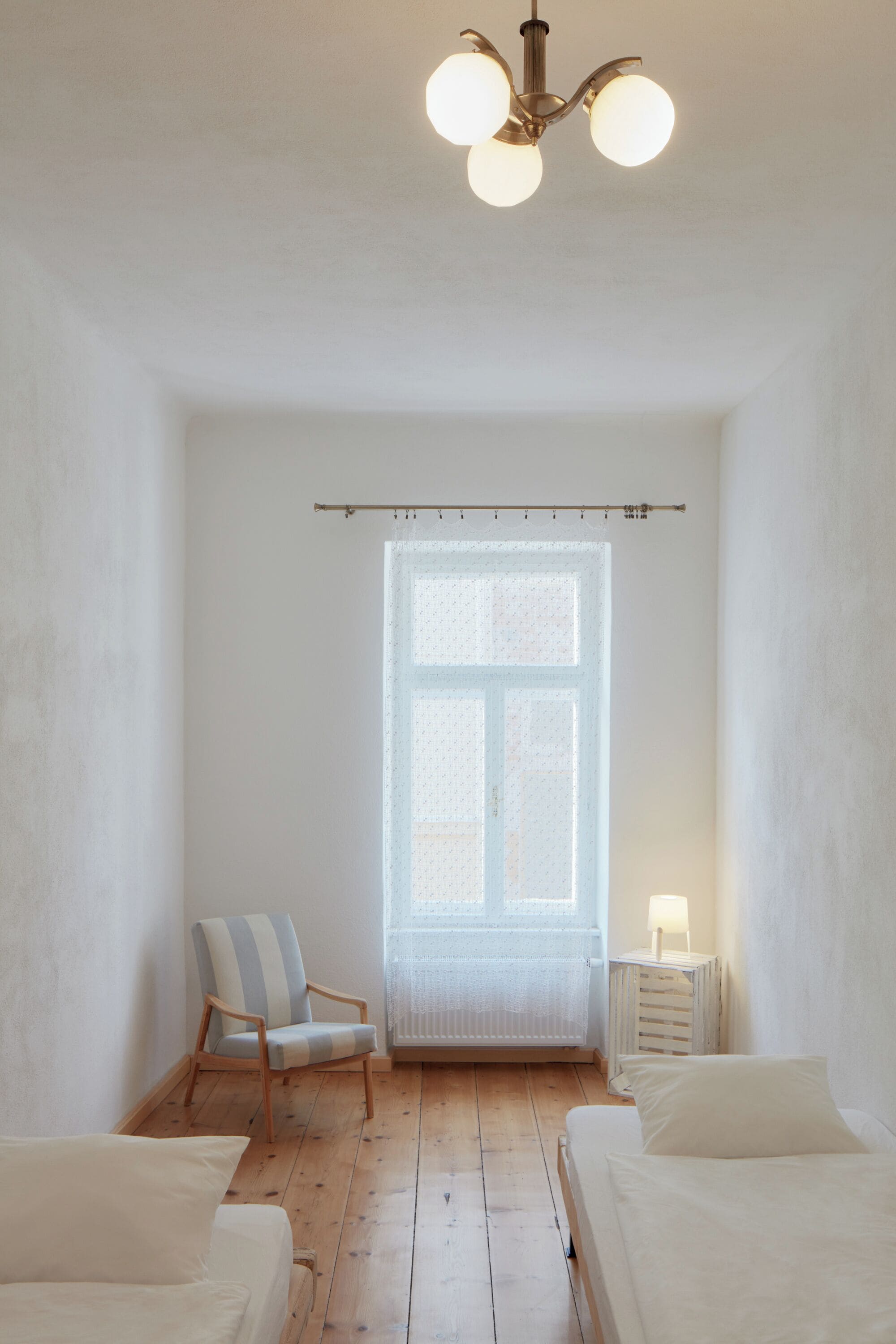 A room with two beds, a chair, a window with a sheer curtain, a nightstand with a lamp, and a ceiling light fixture.