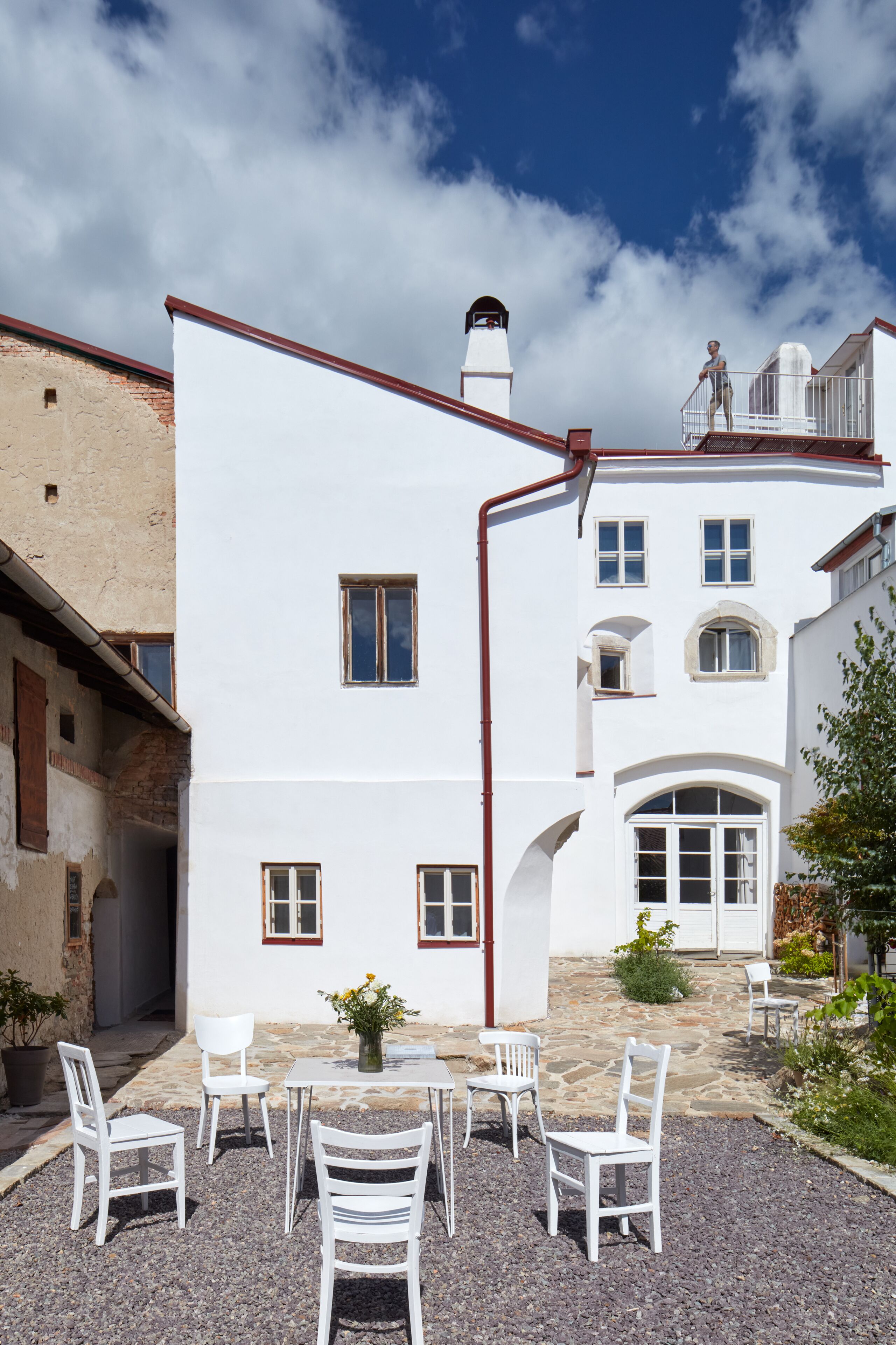 Un bâtiment blanc avec une terrasse en pierre, des chaises blanches et une petite table agrémentée de fleurs.