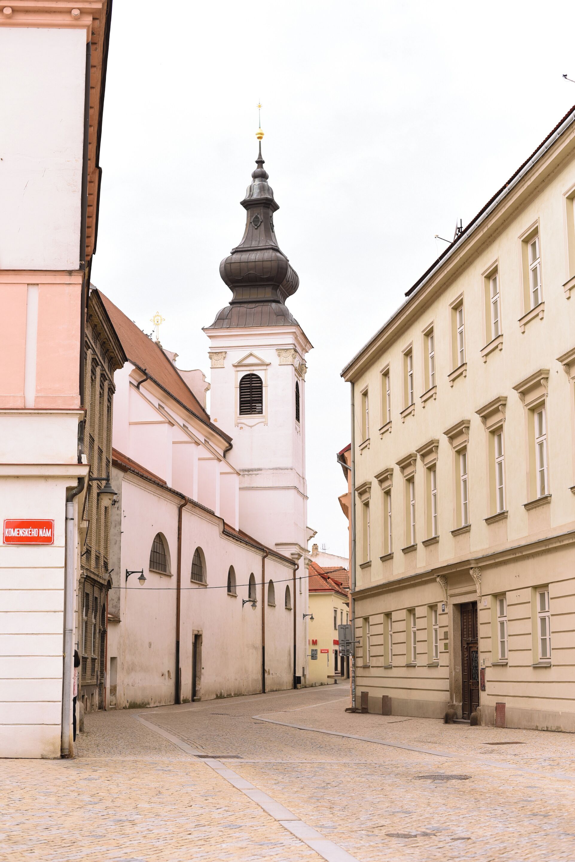 A narrow cobblestone street with historic buildings, including a church with a prominent bell tower.