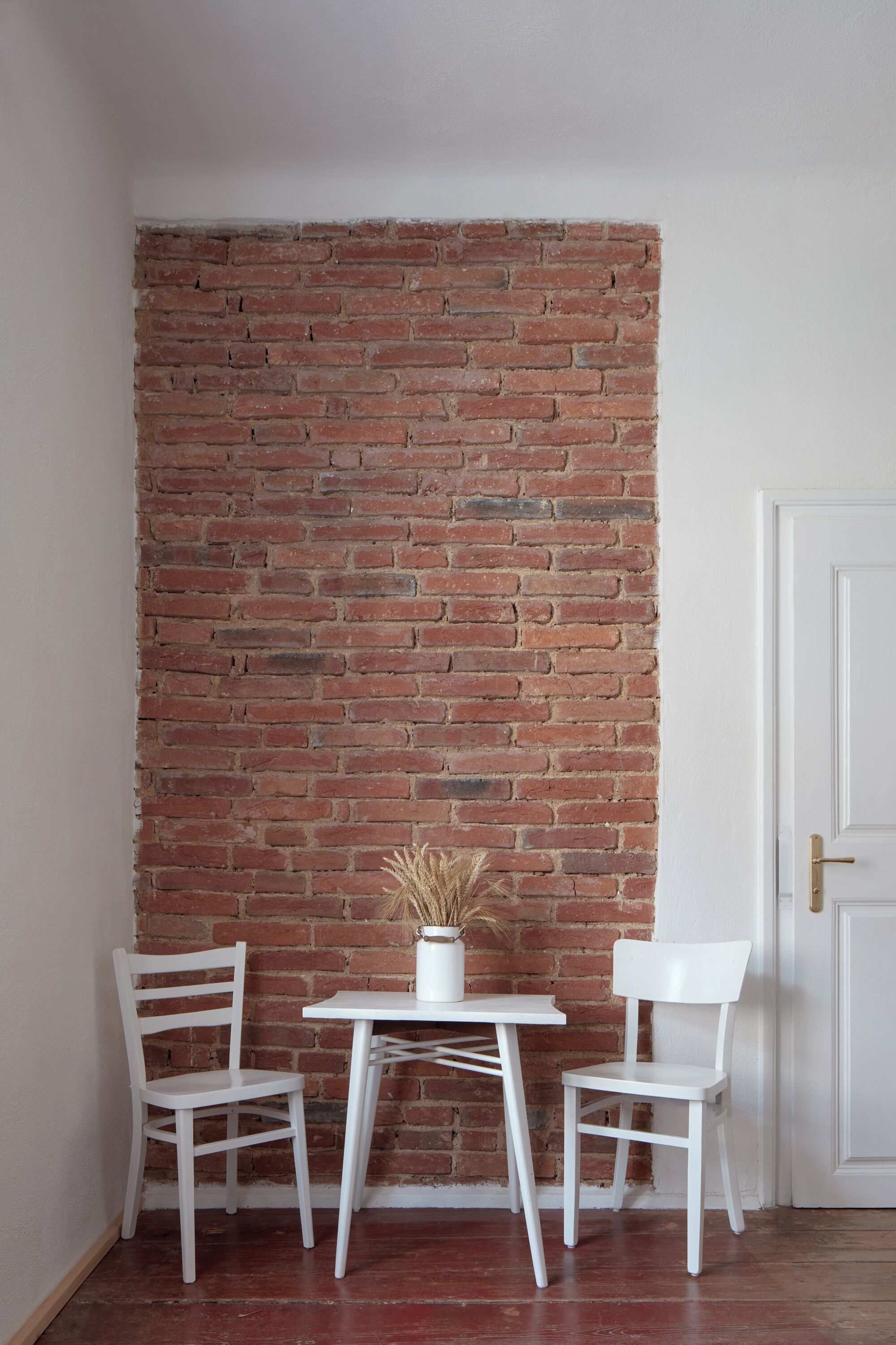 Habitación en esquina con pared de ladrillo, puerta blanca y dos sillas blancas alrededor de una pequeña mesa.