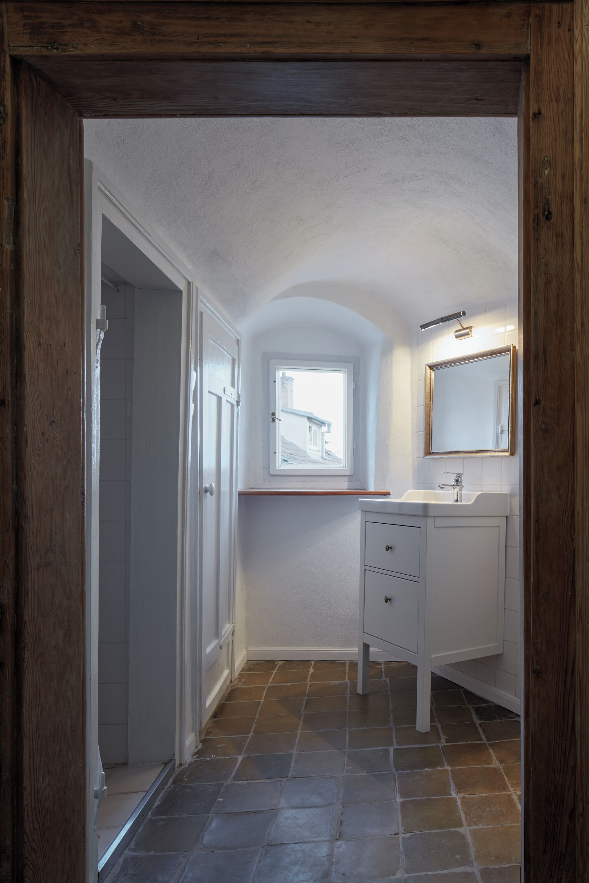 Un couloir étroit mène à une salle de bain équipée d’une fenêtre, d’un lavabo et d’un miroir.