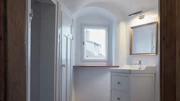 Un couloir étroit mène à une salle de bain équipée d’une fenêtre, d’un lavabo et d’un miroir.