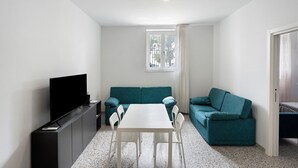 Ein kompakter Wohnbereich mit Flachbildfernseher, einem petrolfarbenen Sofa, einem weißen Esstisch und einer kleinen Küchenzeile.