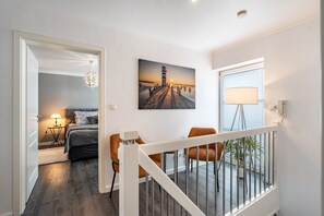 Ein Hotelzimmer mit Bett, Schreibtisch, Stuhl, einem Balkon mit Pflanzen und einem Leuchtturm-Gemälde an der Wand.