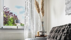 Ett rum med ett fönster, en lampa, en vas med pampasgräs, ett ljus och en fiskstatyett på ett litet bord.