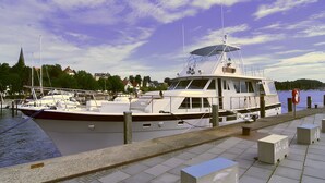 A docked boat with a cabin, a covered area, and a view of the water and buildings in the background.