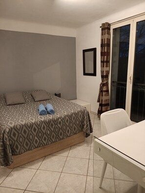 Un dormitorio con una cama, un escritorio, una silla, un ventanal con cortinas y una puerta que da a un balcón.
