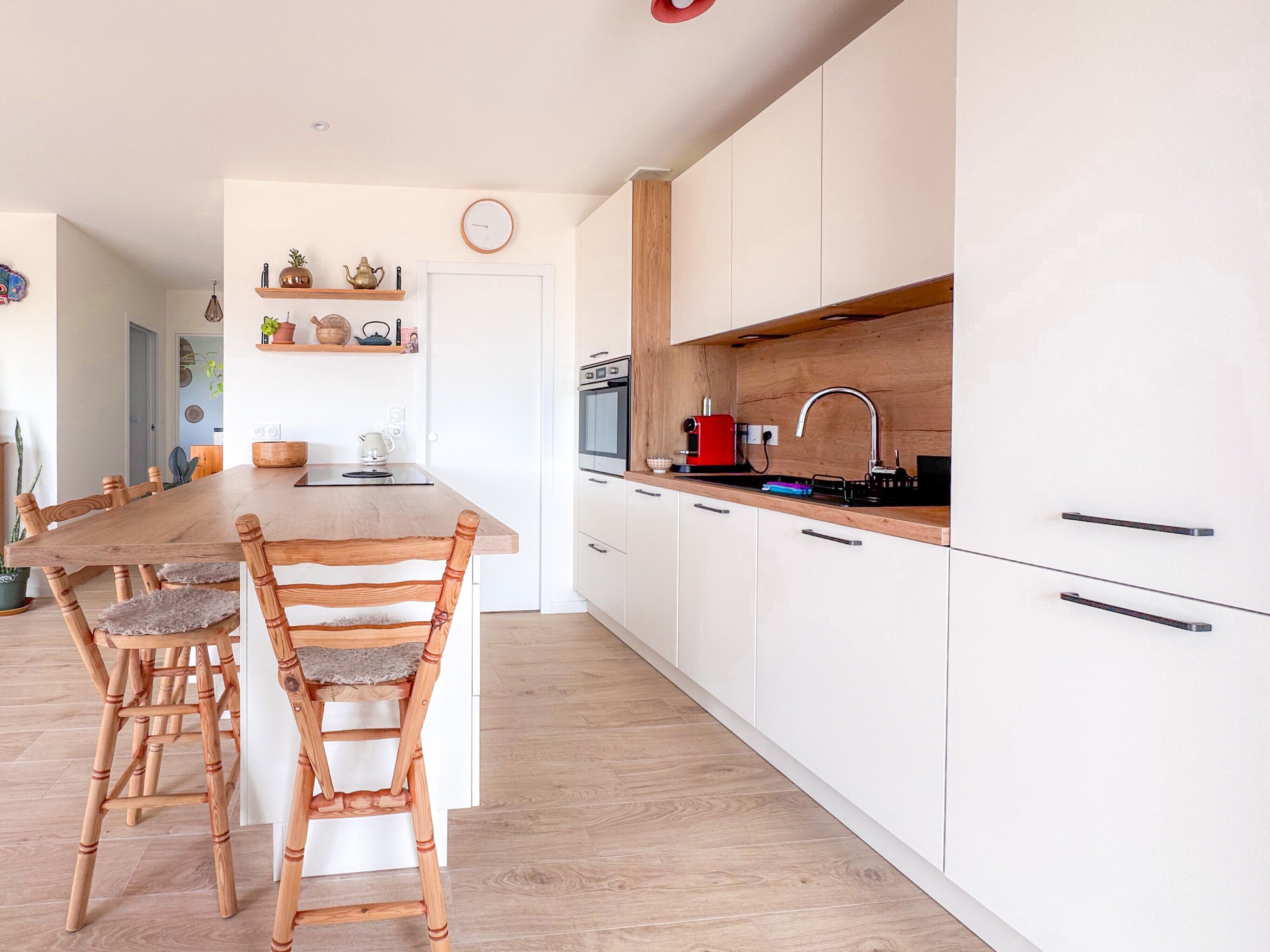 A modern kitchen with wooden chairs, a central island, white cabinets, and a red coffee machine.