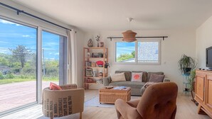 Un salón amplio con un gran ventanal corredera, una estantería de madera, un sofá confortable, un sillón de mimbre y un televisor sobre un mueble de madera.