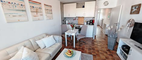 Une cuisine moderne avec des meubles blancs, une table à manger avec des chaises, une machine à café et un téléviseur dans un coin salon agrémenté d’un canapé et d’une table basse.