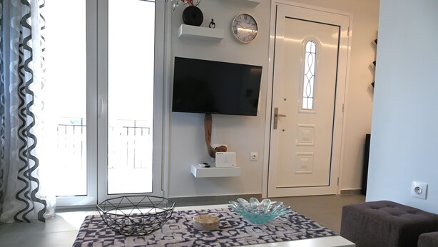 Ein modernes Wohnzimmer mit einem an der Wand montierten Flachbildfernseher, einem gemusterten Teppich, einem kleinen Tisch mit Dekoration und einer Tür mit Fenster.