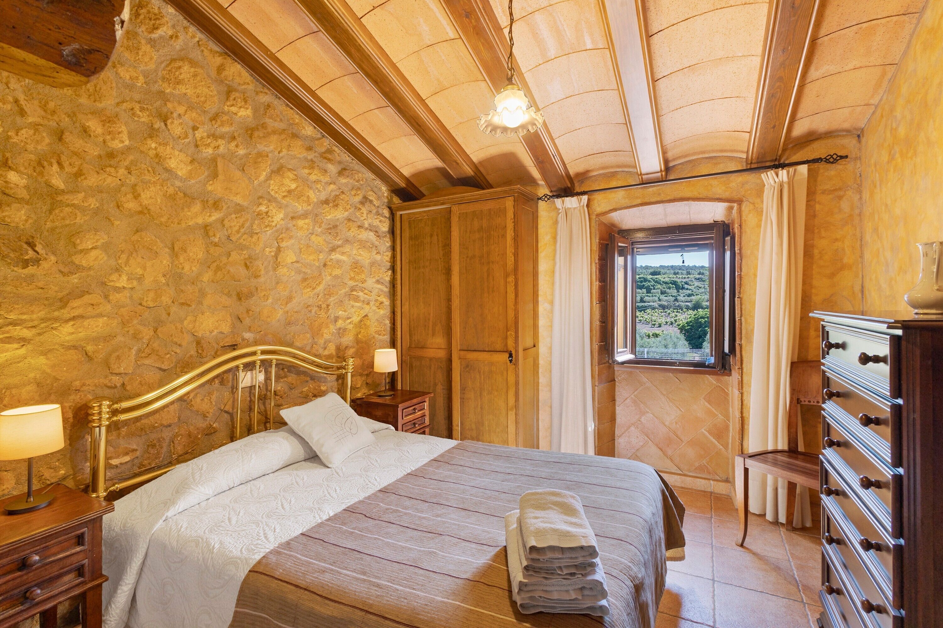 Une chambre avec un mur en pierre, des poutres apparentes en bois au plafond, un lit avec une tête de lit, une table de chevet, une commode et une fenêtre donnant sur un paysage agréable.