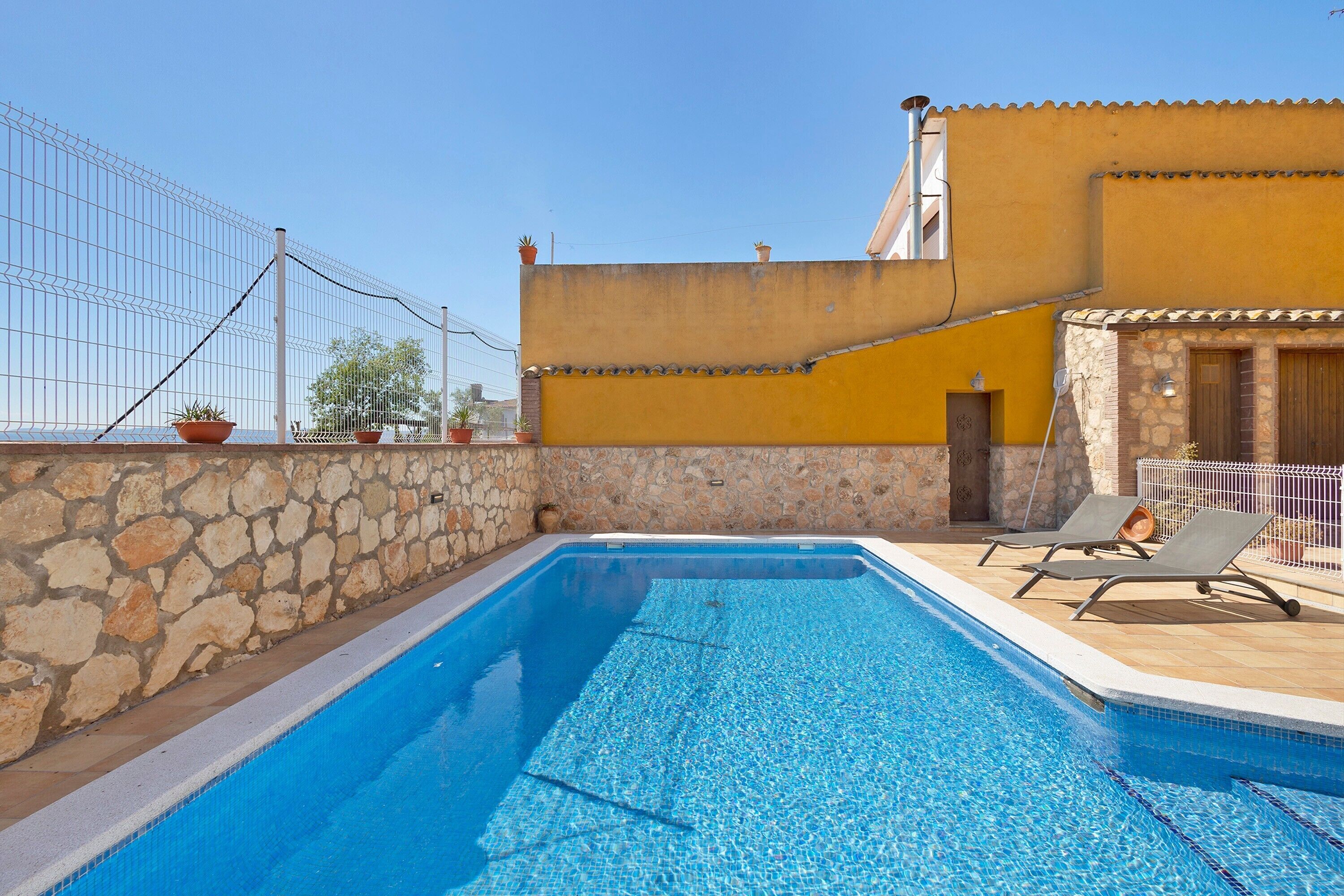 Una piscina con sillas de descanso, un muro de piedra y un edificio amarillo con techo de tejas.