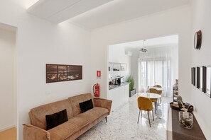 Ein modernes Wohnzimmer mit einer Couch, einem Essbereich mit Stühlen, einer Küche im Hintergrund und einem Feuerlöscher an der Wand.
