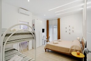A modern hotel room with a bunk bed, a desk, a chair, a bed with a yellow side table, and a window with curtains. - Le Stanze In Centro – Quadruple Room (Naples)