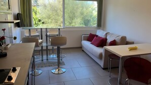 Un salon moderne avec un canapé, une table à manger et des chaises. On y trouve une fenêtre avec des rideaux, un coin cuisine avec une cuisinière, et une petite table sur laquelle est posé un vase de fleurs.