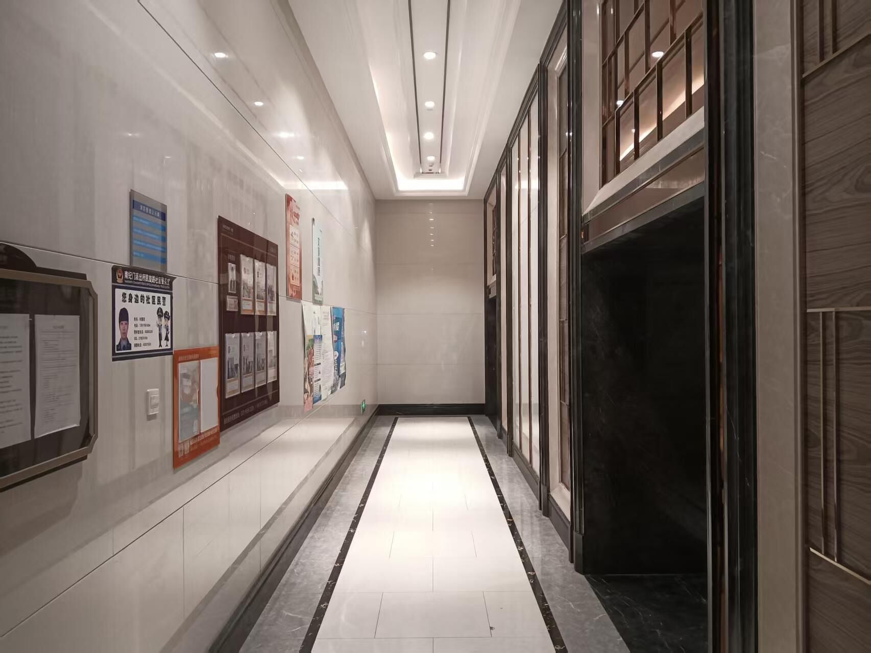A corridor with a tiled floor, a glass elevator, and informational posters on the walls.