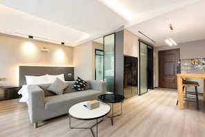 A modern living room with a sofa, a round coffee table, a dining area with a wooden table and chairs, and a sliding glass door leading to a shower area. - J&H-Invis-High-Rise Panoramic Art Hotel (Chongqing)