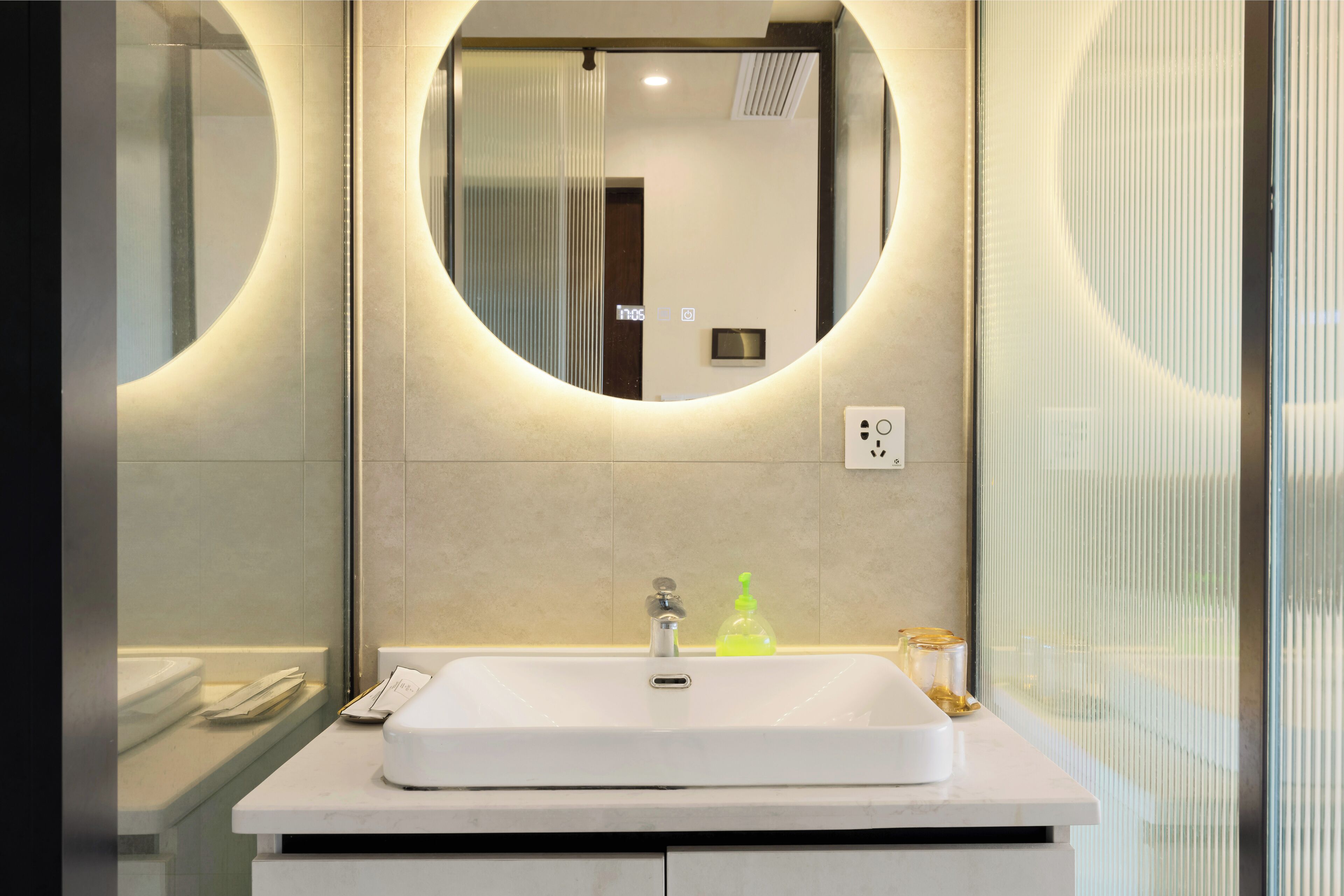 Un baño moderno con un lavamanos blanco, un espejo redondo y una cabina de ducha de vidrio.