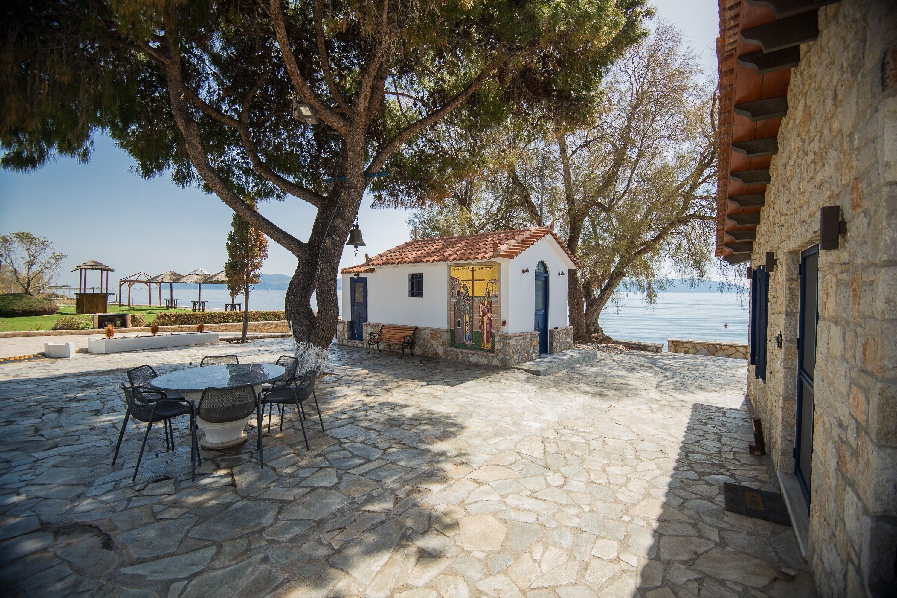 A stone patio with a table and chairs, a small building with a tiled roof, and a view of the sea.
