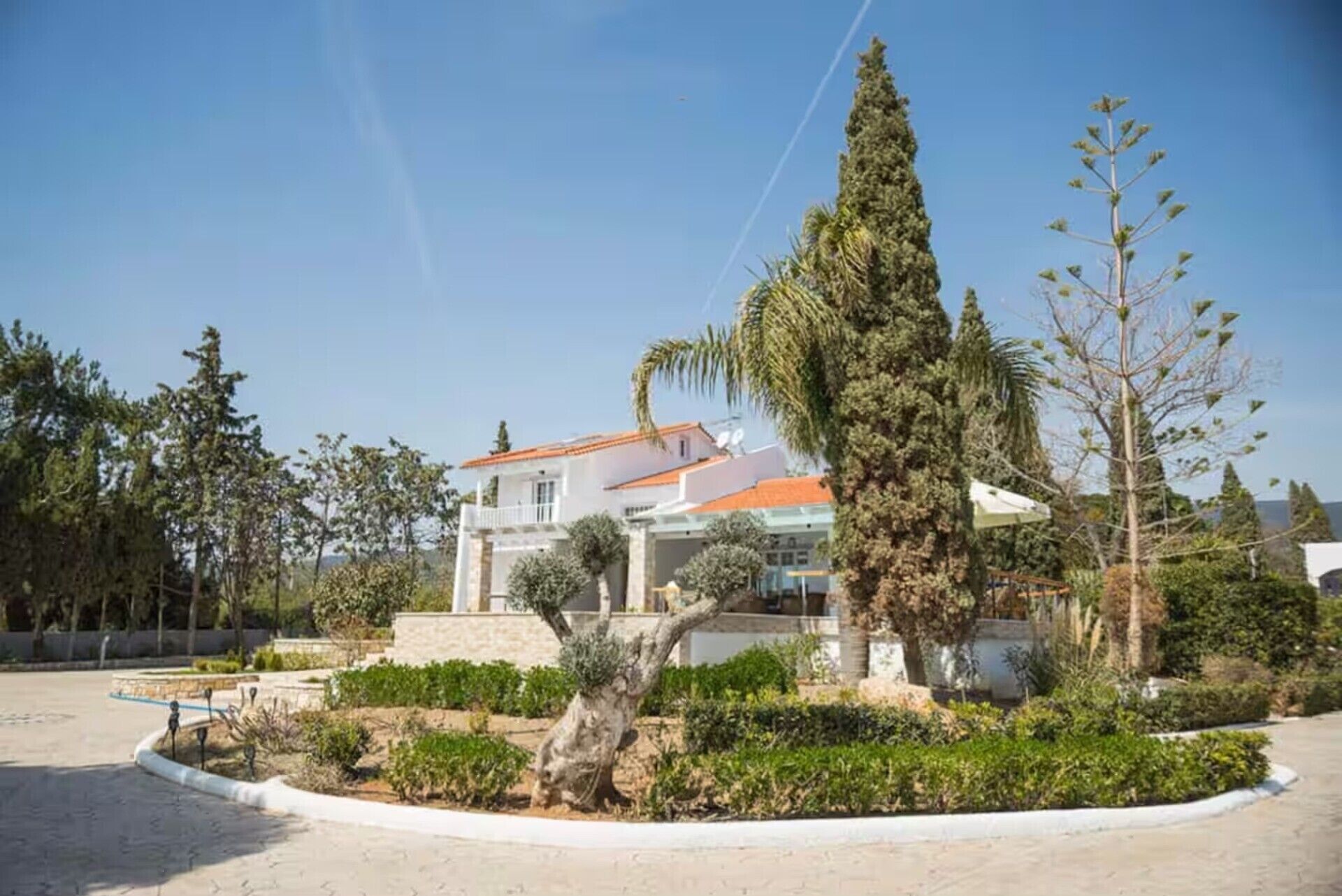 A two-story building with a red-tiled roof, surrounded by a landscaped garden with various shrubs and trees.