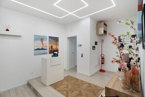 A modern hotel lobby with a reception desk, artwork on the walls, a patterned rug, and a vase with flowers. - Le Stanze In Centro - Deluxe Double Room (Naples)