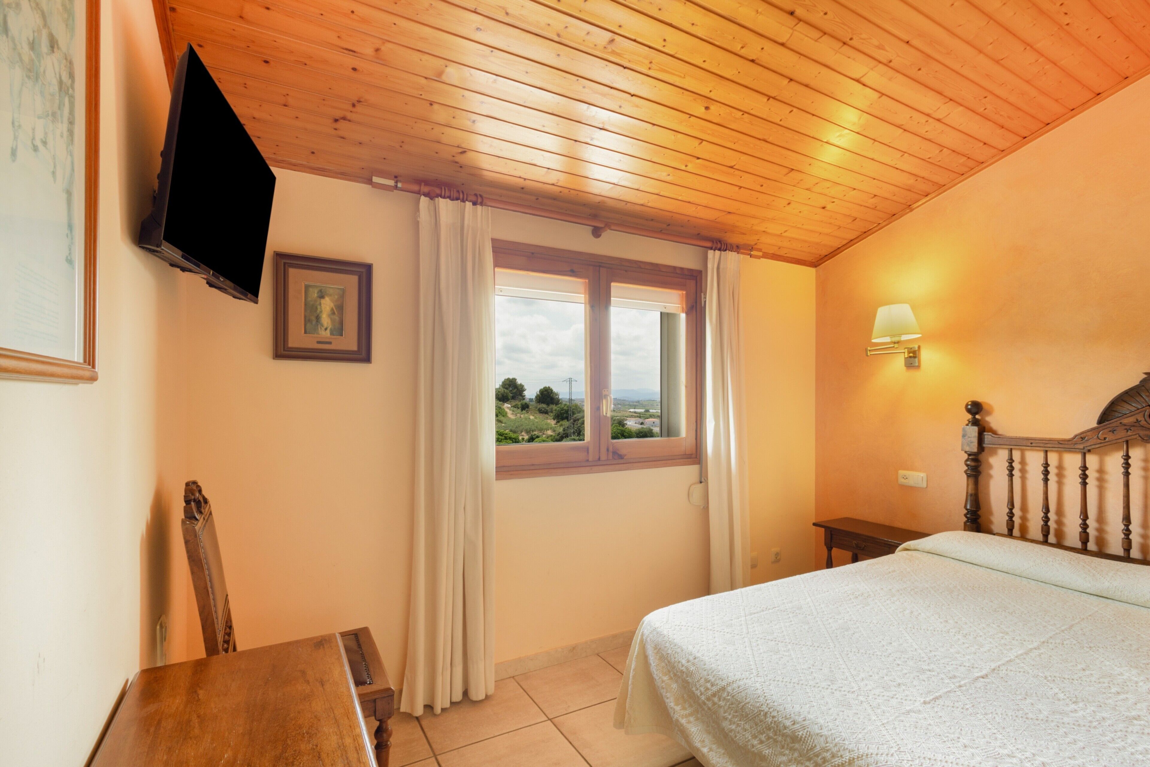 Een slaapkamer met een houten plafond, een bed, een raam met gordijnen, een bedlampje, een aan de muur bevestigde televisie en een lijst met een afbeelding.