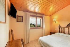 Un dormitorio con techo de madera, una cama, una ventana con cortinas, una lámpara de noche, un televisor montado en la pared y un cuadro enmarcado.