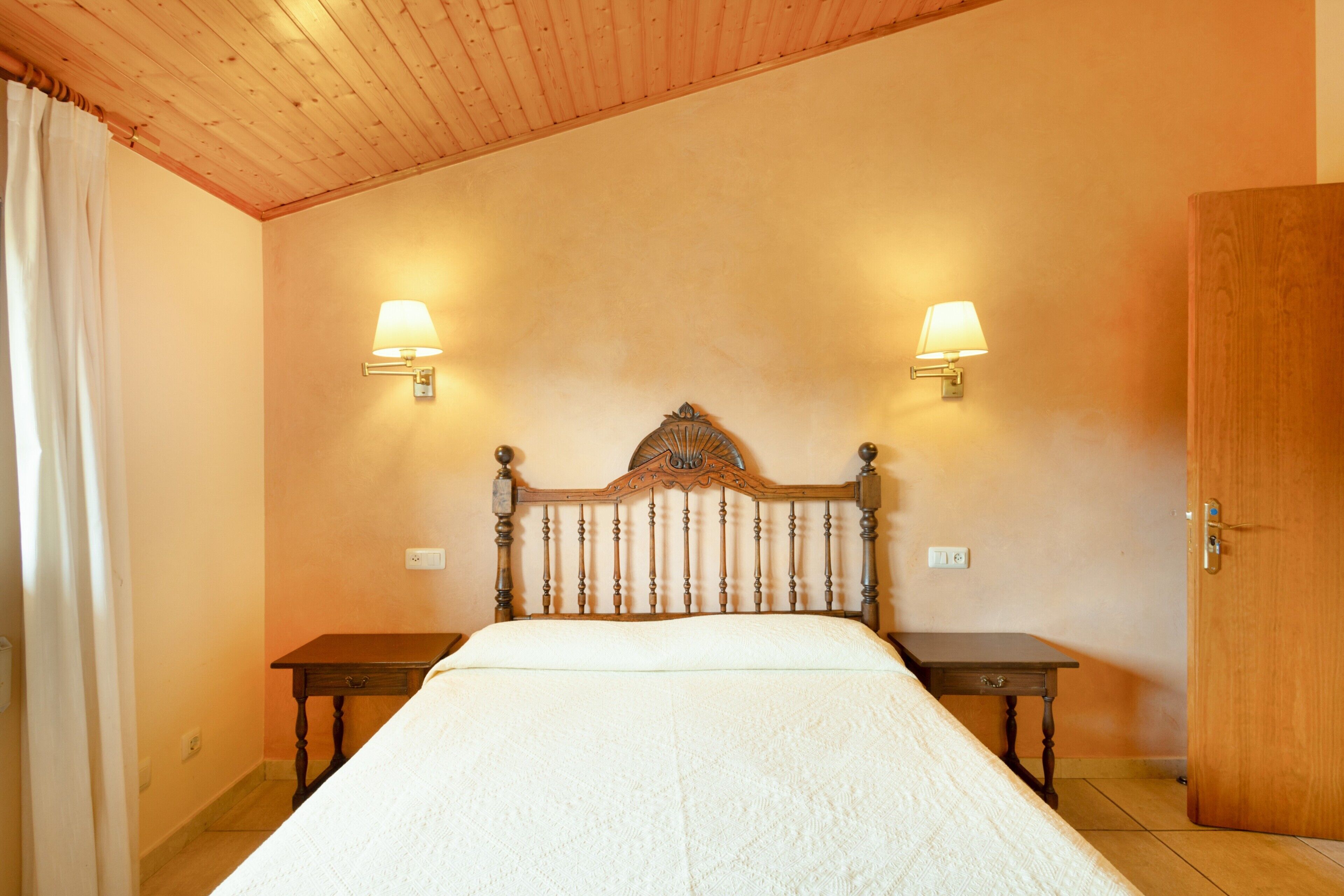 A bedroom with a wooden headboard, two bedside tables, a bed with a white bedspread, two wall-mounted lamps, and a wooden door.